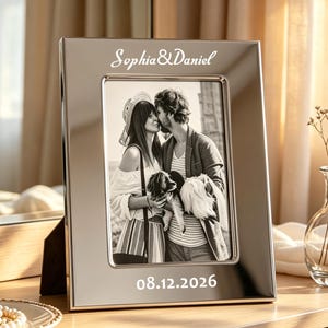 Op de afbeelding: Een rechthoekige fotolijst van zilverkleurig metaal met de gegraveerde tekst Sophia & Daniel aan de bovenkant en 08.12.2026 aan de onderkant. De lijst bevat een zwart-witfoto van twee personen die een kleine hond vasthouden buiten.