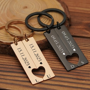 May include: Two personalized keychains with dates and initials. One is gold-colored, the other black. Each keychain has a rectangular tag with a heart-shaped cutout. The dates "15.11.2025" and "09.03.2025" are engraved.