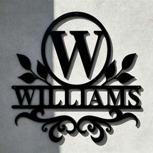 Pode incluir: Uma placa de parede em metal preto com uma letra mai&uacute;scula W grande centralizada dentro de uma moldura circular. Abaixo do c&iacute;rculo, o nome WILLIAMS &eacute; exibido em letras mai&uacute;sculas em negrito, ladeado por motivos de folhas e volutas.