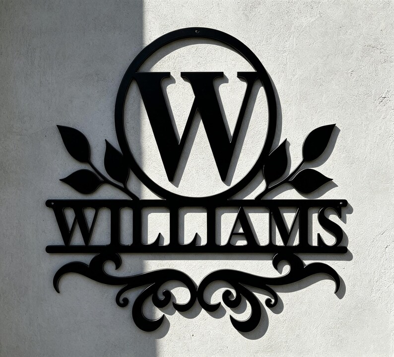 Pode incluir: Uma placa de parede em metal preto com uma letra mai&uacute;scula W grande centralizada dentro de uma moldura circular. Abaixo do c&iacute;rculo, o nome WILLIAMS &eacute; exibido em letras mai&uacute;sculas em negrito, ladeado por motivos de folhas e volutas.
