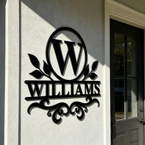 Pode incluir: Uma placa de parede em metal preto com uma moldura circular que envolve a letra W, com o nome WILLIAMS exibido numa barra horizontal abaixo. O design inclui detalhes decorativos de folhas nas laterais e volutas na borda inferior.