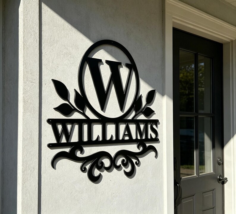 Pode incluir: Uma placa de parede em metal preto com uma moldura circular que envolve a letra W, com o nome WILLIAMS exibido numa barra horizontal abaixo. O design inclui detalhes decorativos de folhas nas laterais e volutas na borda inferior.
