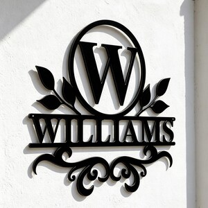 Pode incluir: Uma placa de parede em metal preto com uma letra W grande dentro de um c&iacute;rculo, com o nome WILLIAMS exibido numa barra horizontal abaixo. O design inclui detalhes de folhas decorativas em ambos os lados e volutas ornamentais na base.