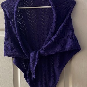 May include: A deep purple triangular shawl featuring an intricate openwork lace pattern throughout the fabric. The knitted garment is draped over a white hanger and tied in a knot at the centre, showcasing the detailed texture of the yarn.