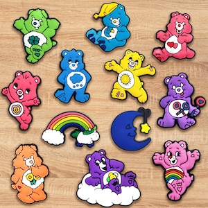 May include: A collection of colorful Care Bear-themed charms. The charms feature various bears in shades of green, blue, pink, yellow, orange, and purple. Other charms include a rainbow, a crescent moon with a star, and a cloud.
