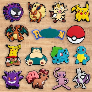 May include: A collection of colorful, cartoon-style character charms, including Pikachu, Eevee, and others. The charms are in various shapes and colors, with a wooden background. The charms are likely for shoes or accessories.