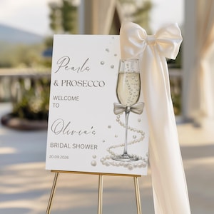 May include: A white sign on a gold easel with the text "Pearls & Prosecco Welcome to Olivia's Bridal Shower 20.09.2026." A champagne glass with a bow and pearl accents is featured. A large cream-colored bow and fabric drape the side.
