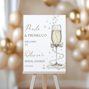 May include: A white sign with the text "Pearls & Prosecco Welcome to Olivia's Bridal Shower 20.09.2026." A champagne glass with a bow and pearl accents is featured. The sign is on an easel with a blurred background of balloons.