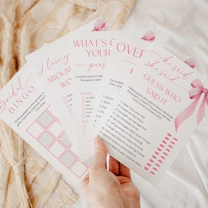 Könnte beinhalten: Eine Sammlung von rosa-weißen Brautparty-Spielkarten. Die Karten enthalten Spiele wie "Bridal Bingo", "I Love You Around the World", "What's in Your Purse?" und "He Said She Said Guess Who Said It". Die Karten werden in einer Hand gehalten.