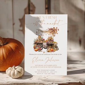 May include: An autumnal invitation card featuring the text "SHE'S FRESH off the market" and a watercolour illustration of a market cart laden with pumpkins, flowers, and produce. A large orange pumpkin and a small white pumpkin are in the foreground.