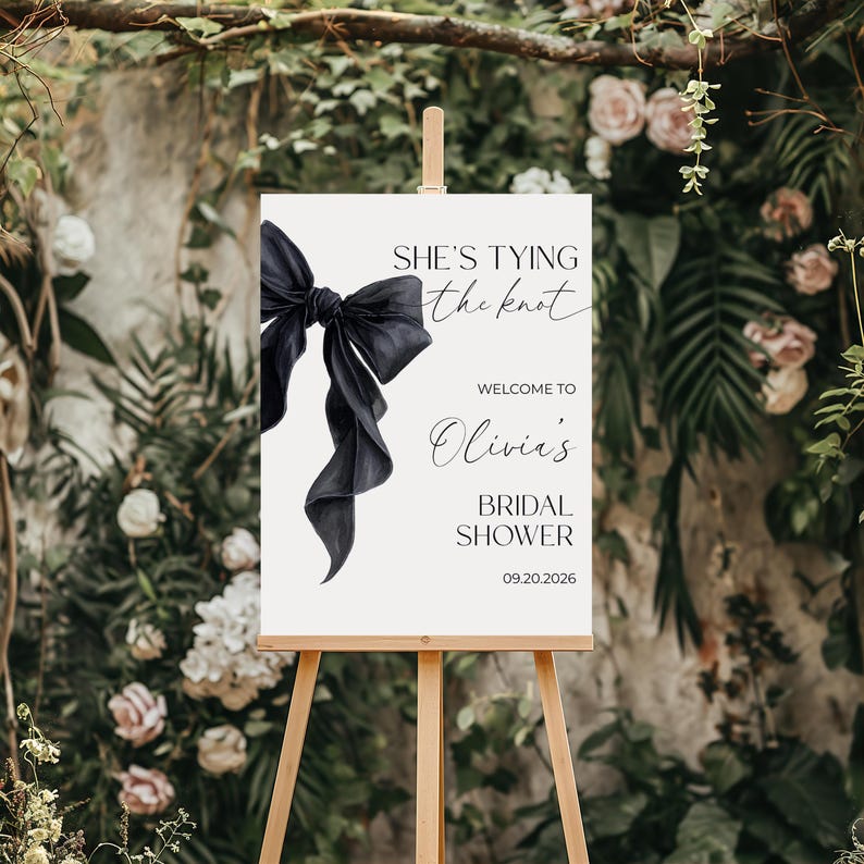 May include: A white sign on a wooden easel with the text "SHE'S TYING the knot" and "WELCOME TO Olivia's BRIDAL SHOWER 09.20.2026". A large black bow is on the left side of the sign. The background is a floral arrangement.