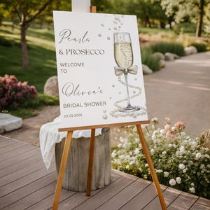 May include: A white sign on a wooden easel with the text "Pearls & Prosecco Welcome to Olivia's Bridal Shower 20.09.2026". The sign features a champagne glass illustration with pearls and a bow.