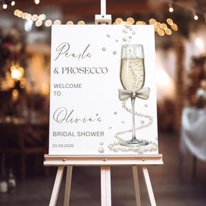 May include: A white sign on an easel with the text "Pearls & Prosecco" and "Welcome to Olivia's Bridal Shower 20.09.2026". A champagne glass with a bow and pearl accents is illustrated on the sign.