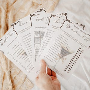 May include: A hand holds a stack of white paper game cards with black text and decorative bow accents. The cards feature bridal shower game prompts like "Would The Bride" and "He Said, She Said". The background is a soft, neutral fabric.