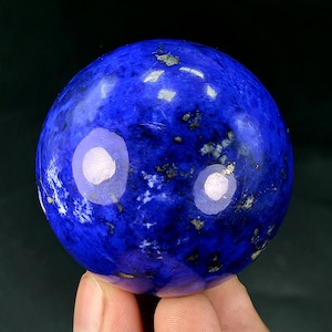 May include: A polished lapis lazuli sphere featuring a deep royal blue colour with natural white calcite marbling and scattered metallic gold pyrite inclusions throughout the smooth, rounded surface of the stone mineral specimen.