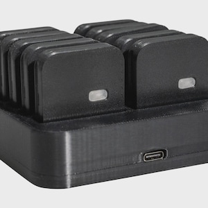 May include: A black charging station with a rectangular base and two sections, each holding multiple rectangular devices. Each device has a small, illuminated indicator. A USB-C port is visible on the front.