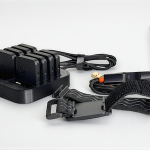 May include: A black charging station with six slots, a USB cable, a wrist strap, and an antenna. The charging station has a sleek, modern design. The USB cable has an orange accent. The wrist strap is black with a velcro closure.