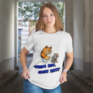 以下が含まれることがあります： 白のTシャツ。漫画の猫とネズミのイラスト。オレンジ色の猫は怒っており、灰色のネズミはロッキングチェアに座っています。Tシャツのテキストは「SWIPE LEFT, GIANT KITTY」です。