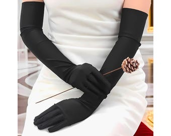 May include: Black opera gloves, extending to the upper arm, are featured in this image. The gloves are made of a smooth, opaque material. A brown pinecone is held between the gloved hands. The background is a white dress.