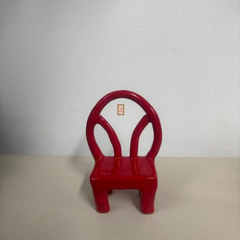May include: A small, bright red chair sculpture with a rounded backrest featuring two open, curved cutouts. The chair has four sturdy legs and a smooth, glossy finish. A small rectangular tag with the number 2 is visible in the center.