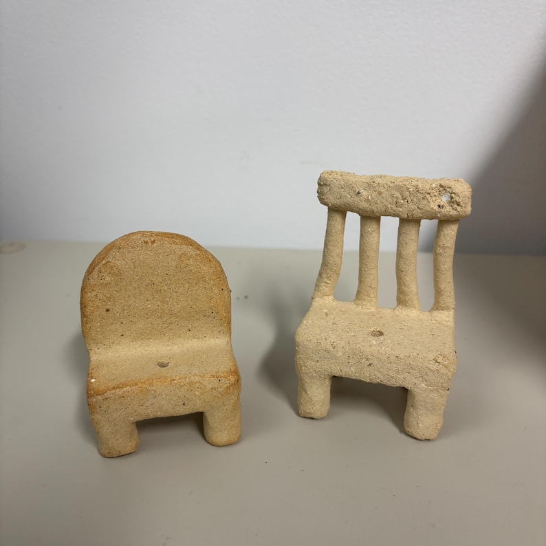 May include: Two miniature chairs crafted from a light beige, textured clay material. One chair features a solid, rounded backrest, while the other chair has a slatted backrest with four vertical supports. Both pieces have four legs.