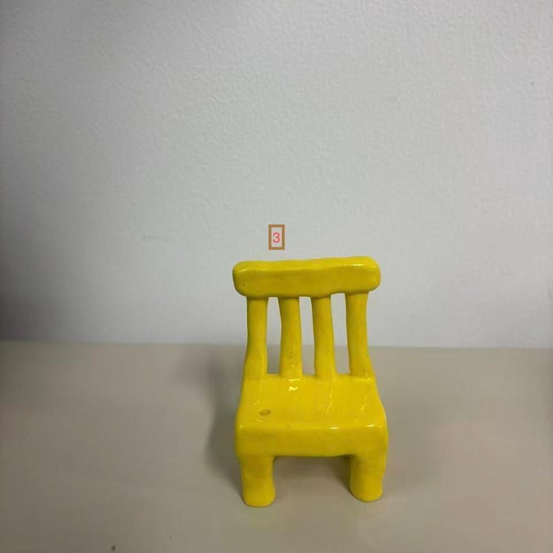 May include: A small, bright yellow chair figurine with a smooth, glossy finish. The chair features a rectangular backrest supported by four vertical slats and a solid seat, with the number 3 enclosed in a small orange box floating above it.