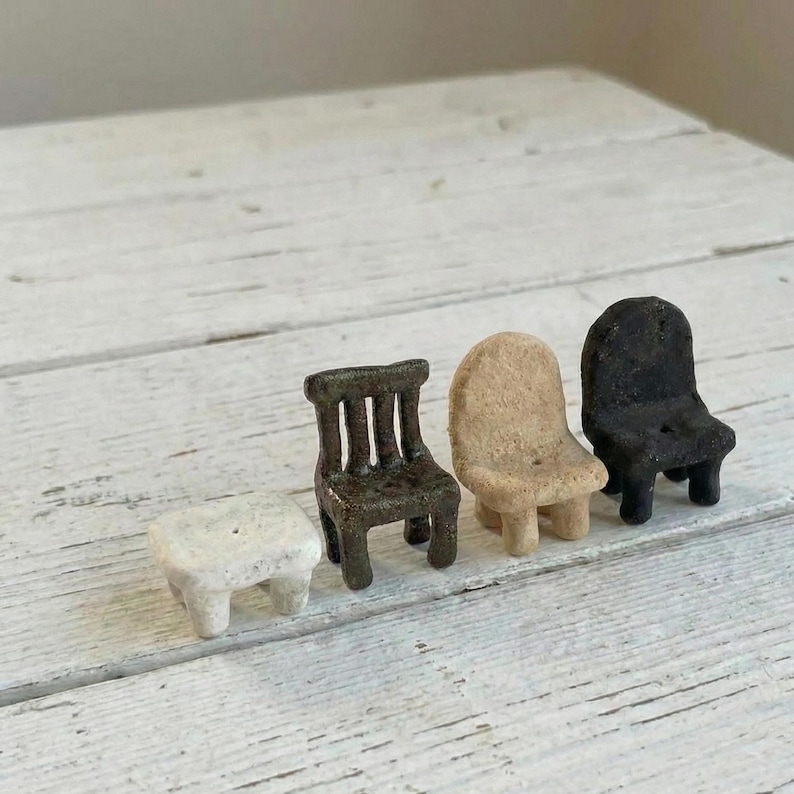 May include: A set of four miniature furniture pieces including a small white rectangular stool, a dark brown chair with a slatted back, a beige rounded chair, and a black rounded chair, all featuring a textured, matte, and rustic finish.