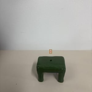 May include: A small, dark green, rectangular stool with four short, thick legs and a single circular hole centered on the flat top surface. The object has a smooth, glossy finish and a simple, solid construction with rounded edges.