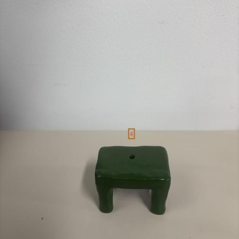 May include: A small, dark green, rectangular stool with four short, thick legs and a single circular hole centered on the flat top surface. The object has a smooth, glossy finish and a simple, solid construction with rounded edges.