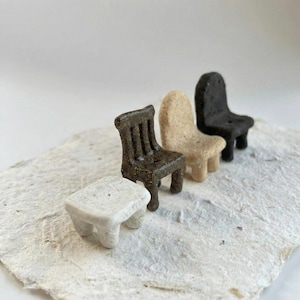 May include: A collection of four miniature furniture pieces made of textured material. The set includes a small white rectangular stool, a dark brown chair with a slatted back, a beige chair with a solid back, and a black chair with a solid back.