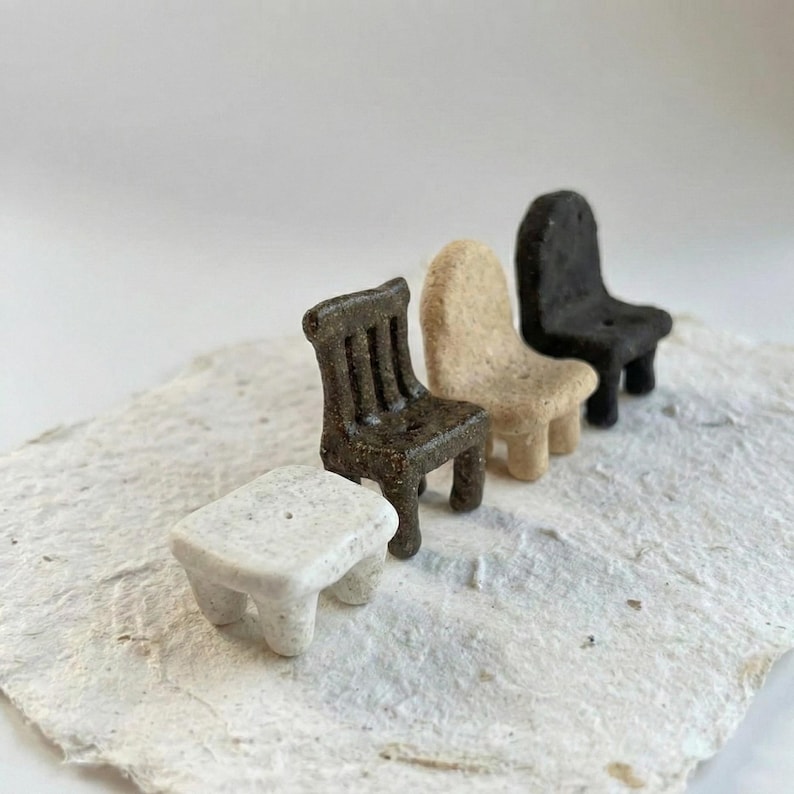 May include: A collection of four miniature furniture pieces made of textured material. The set includes a small white rectangular stool, a dark brown chair with a slatted back, a beige chair with a solid back, and a black chair with a solid back.