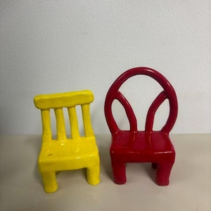 May include: Two small, glossy ceramic chairs. One chair is bright yellow with a rectangular backrest featuring three vertical slats. The other chair is vibrant red with a rounded, arched backrest and a smooth, continuous frame design.