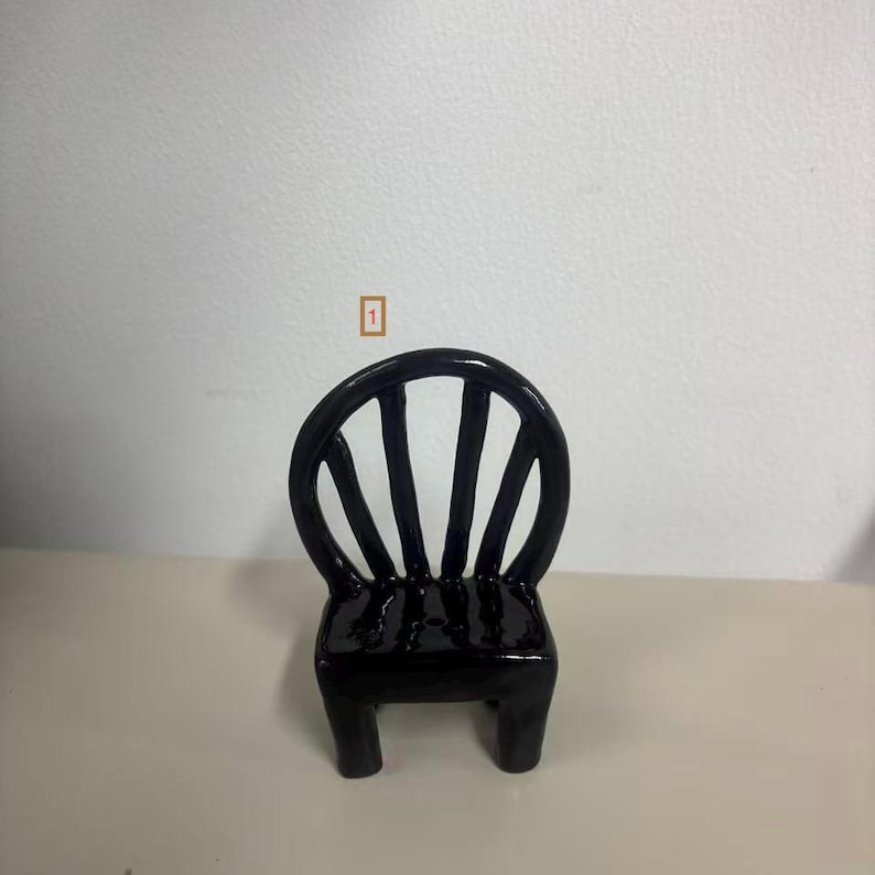 May include: A small, glossy black ceramic chair figurine featuring a rounded backrest with four vertical slats and a square seat. The object has a smooth, reflective finish and is marked with a small orange square containing the number 1.