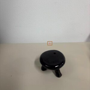 May include: A small, black, three-legged ceramic stool or stand with a smooth, glossy finish and a circular top. The object features a single, small, circular hole near the edge of the top surface and is marked with the number 5 in a small box.