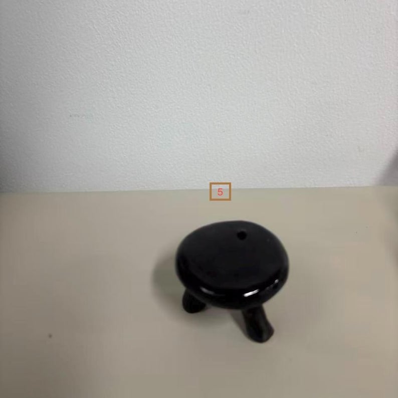 May include: A small, black, three-legged ceramic stool or stand with a smooth, glossy finish and a circular top. The object features a single, small, circular hole near the edge of the top surface and is marked with the number 5 in a small box.