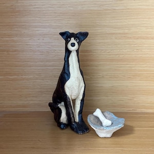 May include: A black and white ceramic dog figurine in a seated position with upright ears and wide eyes. Beside the dog is a small, light-colored ceramic bowl containing a single white bone-shaped object, both crafted with a textured finish.