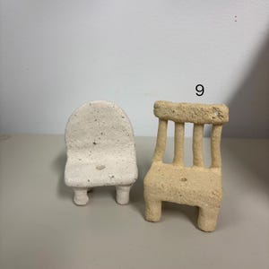 May include: Two miniature chairs made of a textured, porous material. The chair on the left is off-white with a rounded backrest. The chair on the right is tan, features a slatted backrest, and is marked with the number 9 above it.