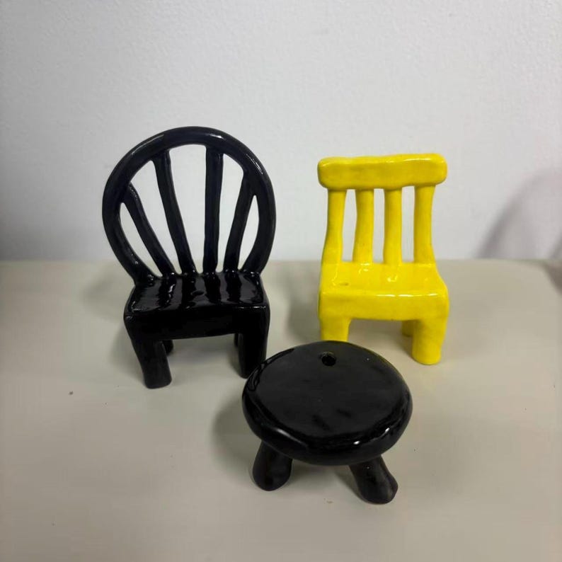 May include: A set of three miniature ceramic furniture pieces consisting of a black chair with a rounded back, a yellow chair with a rectangular back, and a small black round table with three legs, all featuring a smooth, glossy finish.