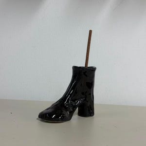 May include: A glossy black ceramic incense holder shaped like a high-heeled ankle boot with a textured, uneven surface. A single brown incense stick is inserted into the top opening of the boot, standing upright against the plain background.