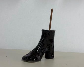 May include: A glossy black ceramic incense holder shaped like a high-heeled ankle boot. The boot features a textured, uneven surface and a block heel. A single brown incense stick is inserted into the top opening of the boot.