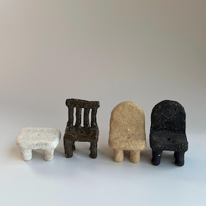 May include: A collection of four miniature furniture pieces with textured surfaces. The set includes a small white rectangular stool, a dark brown chair with a slatted back, a beige chair with a rounded back, and a black chair with a rounded back.