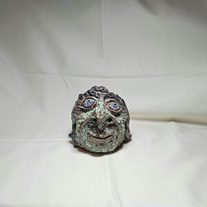 May include: A ceramic sculpture of a face with blue eyes, a smiling mouth, and textured skin. The sculpture has a dark, textured hair and is primarily green and brown in color. The piece is set against a white background.