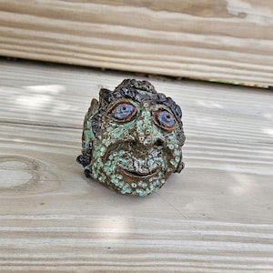 May include: A small, hand-sculpted ceramic head with a textured, mottled surface in shades of mint green, brown, and black. The figure features two prominent, glossy blue eyes, a defined nose, and a subtle, curved mouth, creating a detailed face.