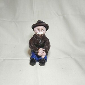 May include: A small ceramic figurine of a person wearing a dark brown hat and matching jacket, blue trousers, and black shoes. The figure has a pale complexion, a prominent white mustache, and is depicted in a seated position with hands clasped.