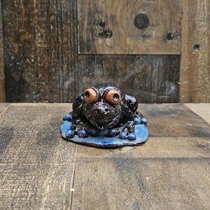 May include: A dark-colored ceramic frog figurine with large, prominent brown eyes and textured skin, resting on a flat, blue-glazed ceramic lily pad base. The frog features distinct toes and a wide mouth, finished with a glossy, mottled glaze.