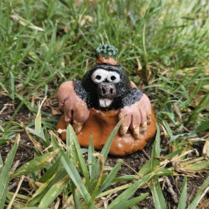 May include: A hand-painted ceramic figurine of a mole emerging from a mound of brown earth. The mole has a black body, large white eyes with black pupils, a pinkish snout, and two prominent white front teeth. A small green textured cap sits atop its head.