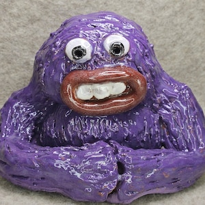 May include: A purple ceramic monster with googly eyes, a wide open mouth with white teeth, and a red tongue. The monster has a furry texture.