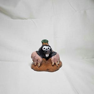 May include: A handcrafted ceramic mole figurine. The mole has a black body, pink paws, and a brown base. It has a green leafy detail on its head and a white face with black eyes. The figurine is a whimsical decorative item.