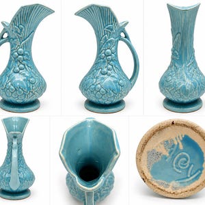 May include: A light blue ceramic pitcher featuring a relief pattern of grapes and leaves on the body, a curved handle, and a flared rim. The base is circular and the underside shows a blue glazed center with a stamped mark and rough edges.