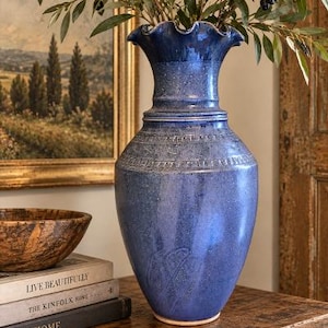 May include: A tall, blue ceramic vase with a ruffled rim and a speckled glaze finish. The vase features a band of embossed geometric patterns around the upper shoulder and a subtle, etched leaf design on the lower body, holding green leafy branches.